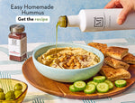 Hummus in a bowl with pita bread and vegetables on a table being topped with Arbequina Extra Virgin Olive Oil and Sumac.