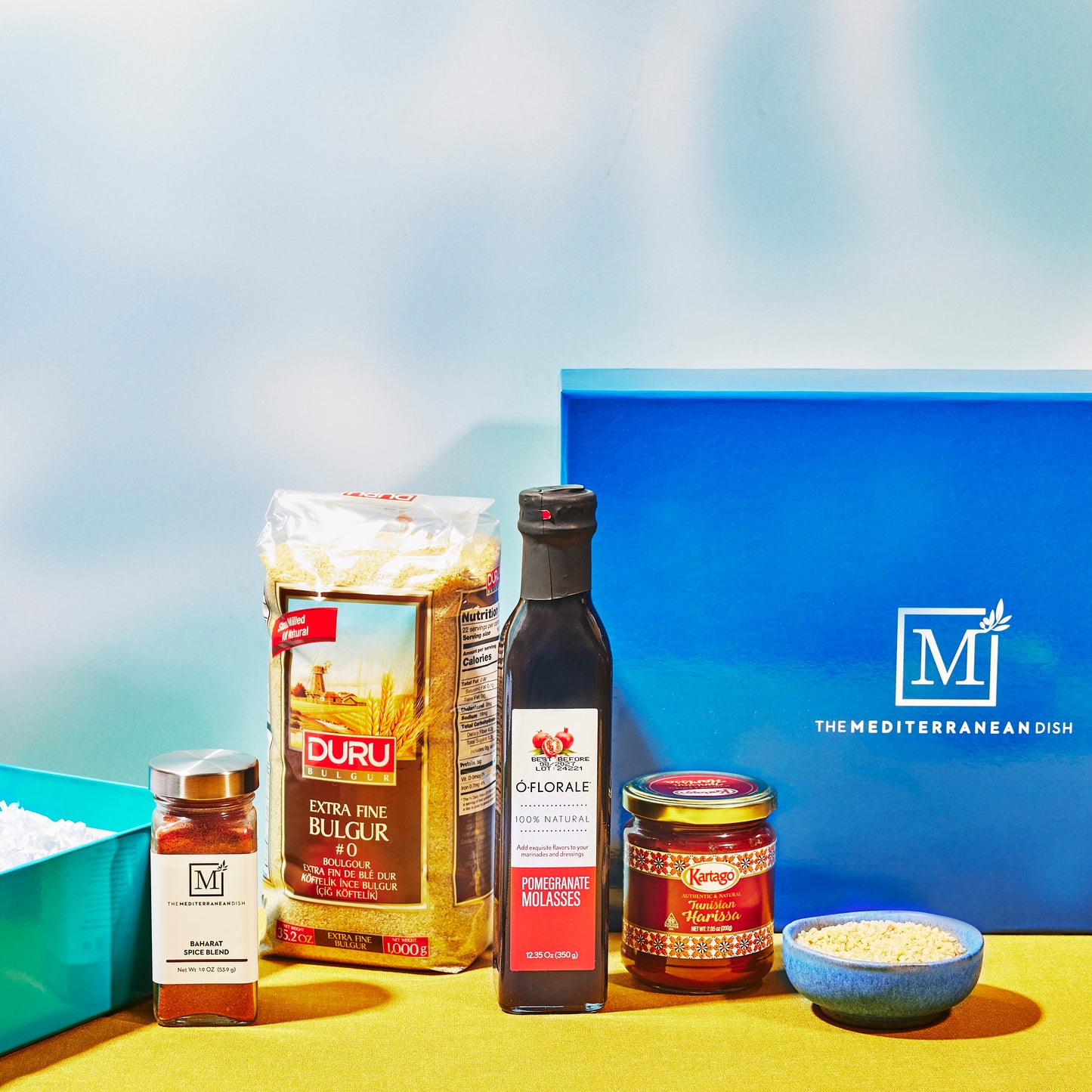 Assorted food products including a bag of durum wheat, olive oil, and other ingredients on a blue and yellow background.