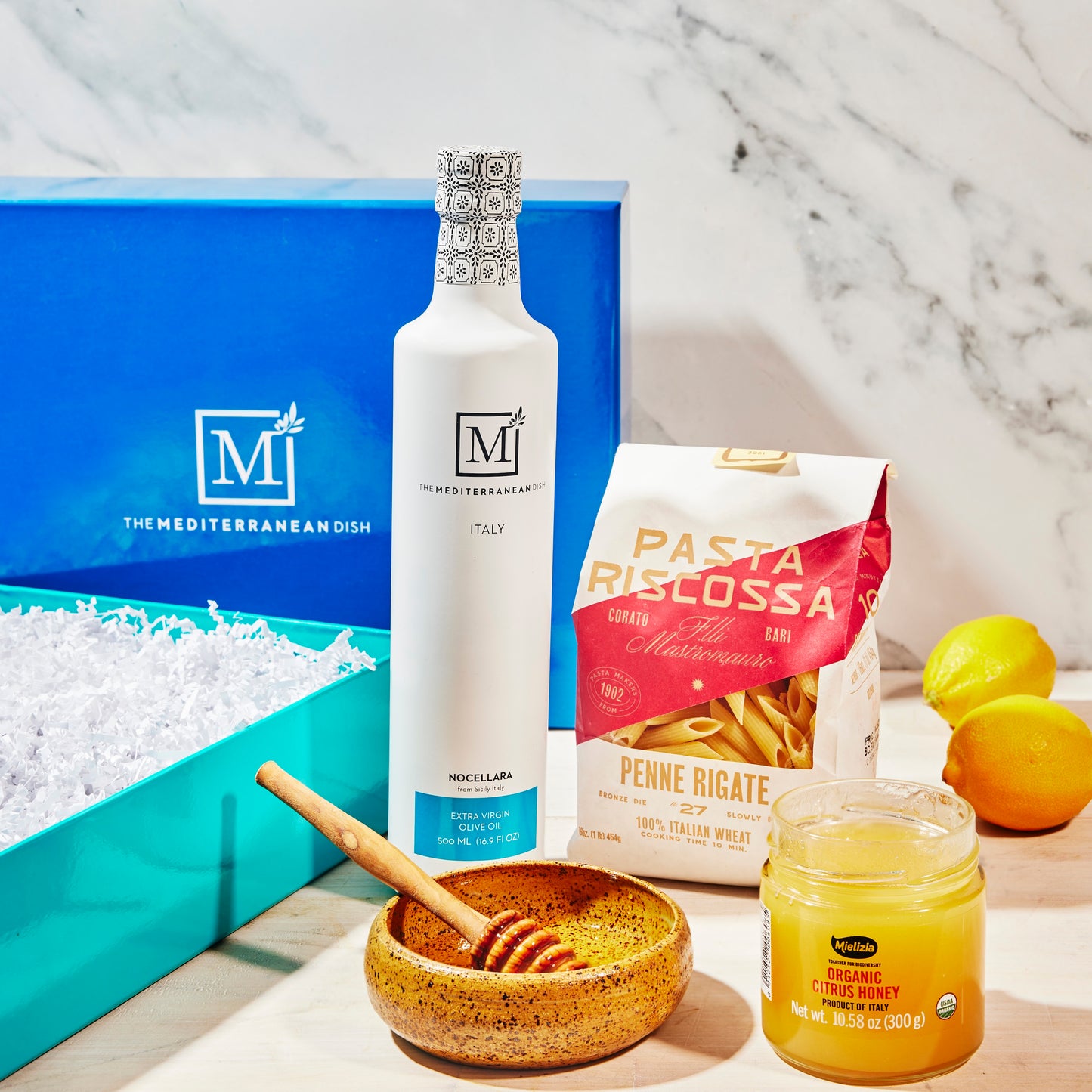 Assorted food items including a bottle, pasta, lemons, and a jar on a marble surface with The Mediterranean Dish branding.