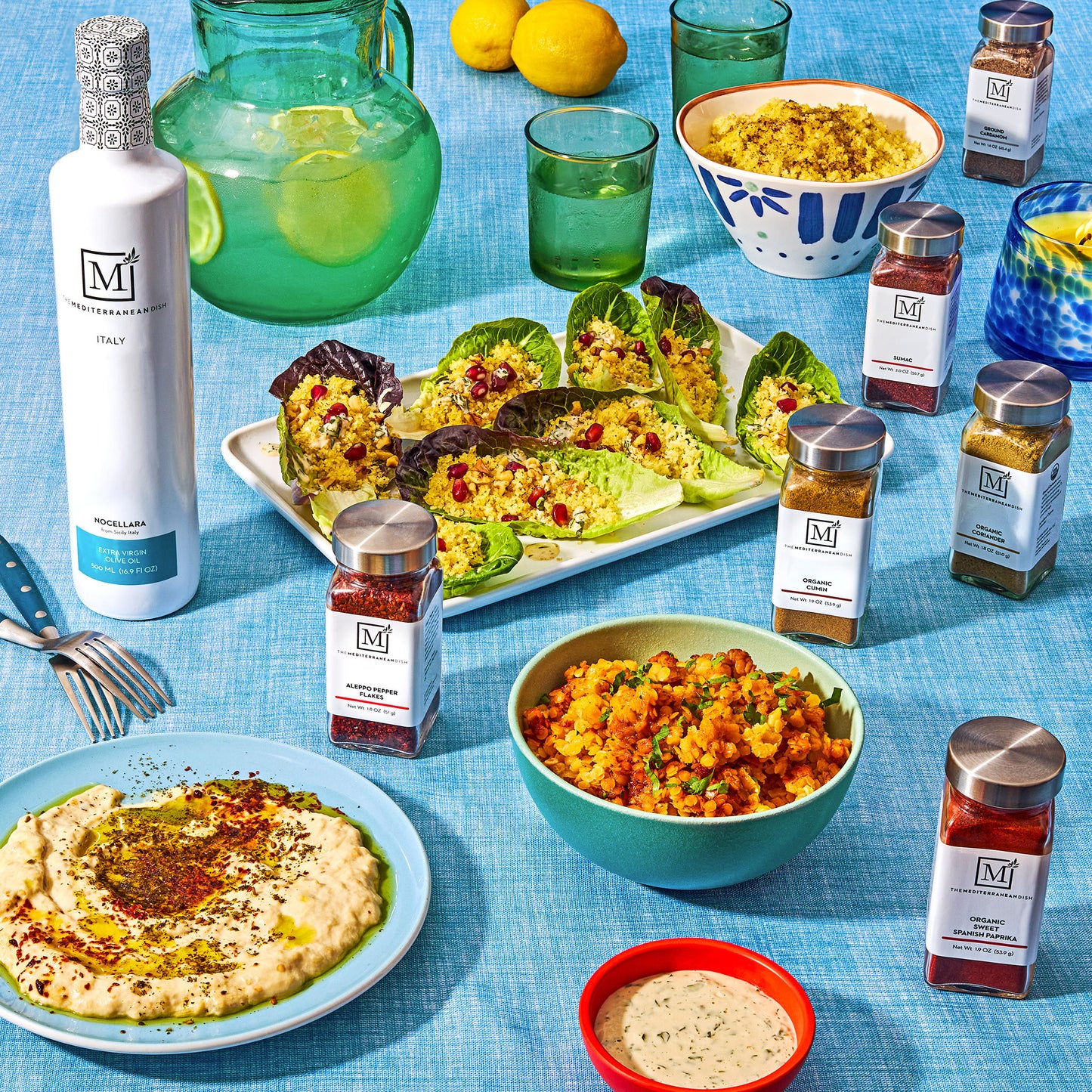 Various food items from The Mediterranean Dish on a blue background with a bowl of hummus, lettuce cups, and lemonade.
