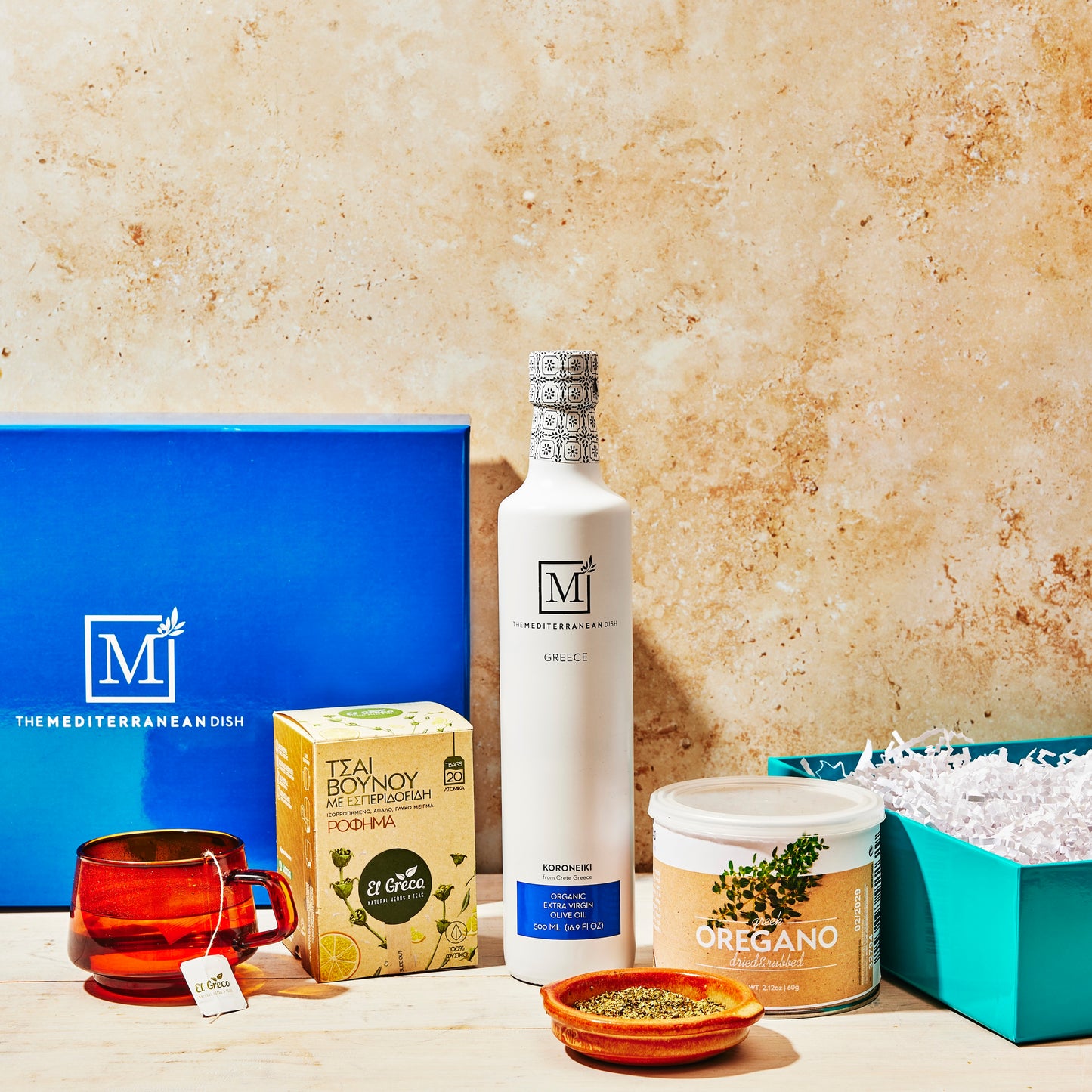 Collection of Mediterranean-themed products including a bottle, tea box, and container on a textured surface.
