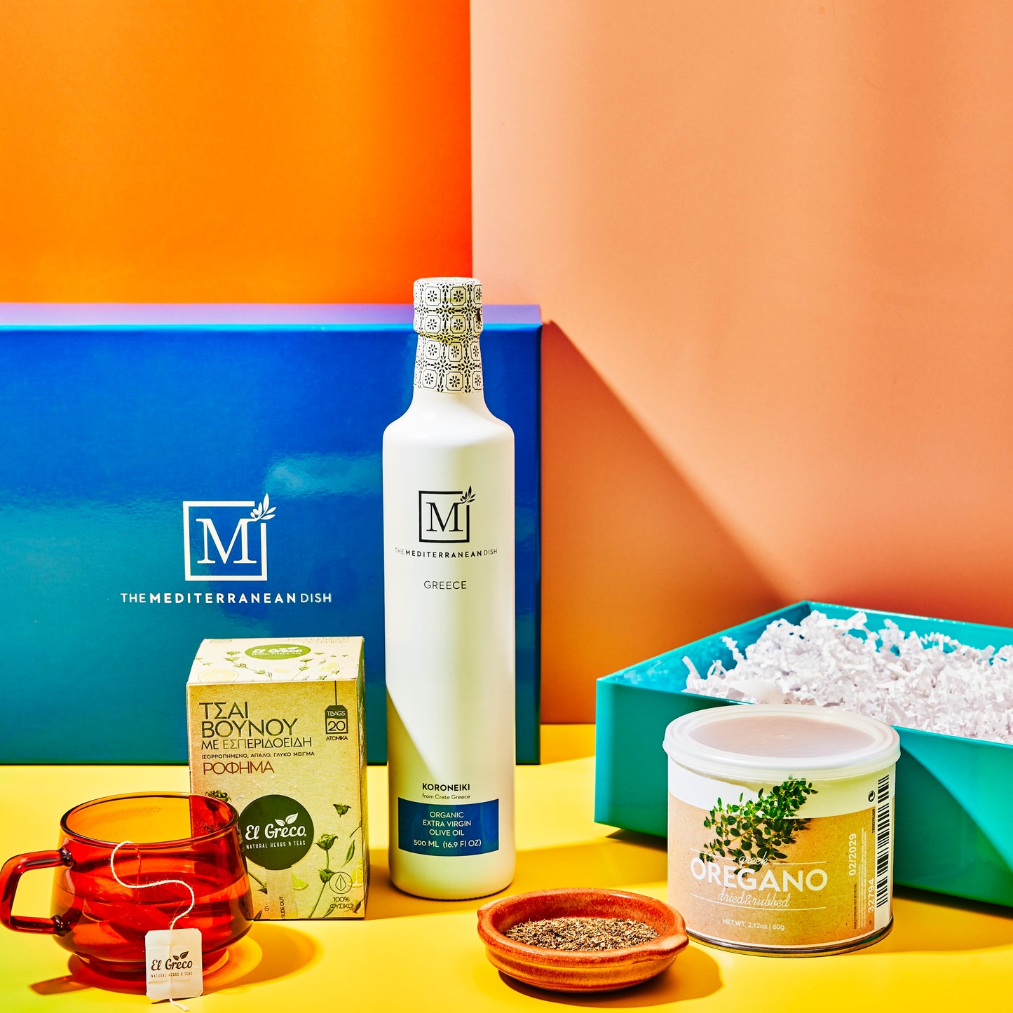Assorted food products including olive oil, tea, and oregano on a colorful background with 'The Mediterranean Dish' branding.
