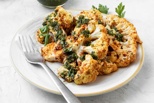 Two cauliflower steaks attractively layered on a plate with one resting on top of the other. Both have been dressed with green Chermoula. There is a small bowl of Chermoula on the side and a metal fork on the plate.