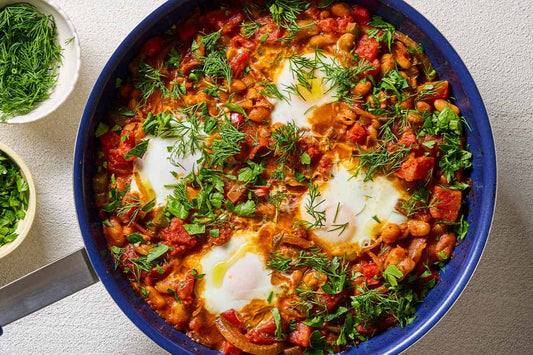 An overhead photo of white bean shakshuka garnished with dill and parsley in a skillet. Next to this are small bowls of dill and parsley.