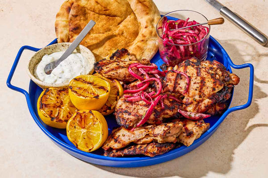 A close up of grilled sumac chicken thighs on a serving platter with pickled red onions, grilled lemon halves, tzatziki sauce, and pita bread.