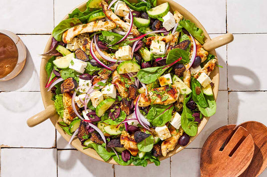 An overhead photo of Greek-stye chicken spinach salad in a serving bowl next to wooden serving utensils and a glass of the dressing.