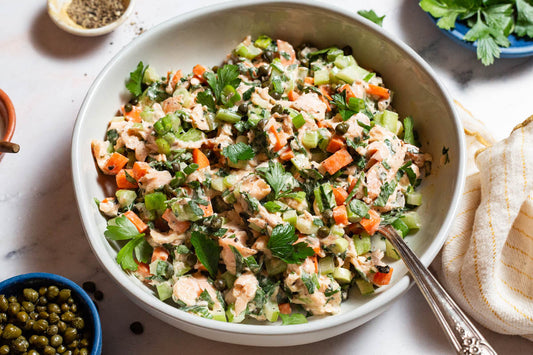 A overhead close up photo of salmon salad in a serving bowl with a spoon next to bowls of capers and parsley.