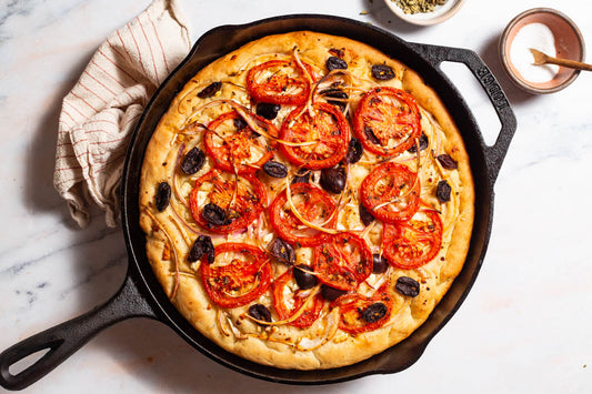 An entire sliced Greek pizza on a wooden cutting board next to bowls of oregano and kalamata olives.