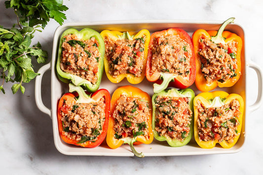 An overhead photo of 8 cooked chicken stuffed peppers in a baking dish with a serving spoon. Surrounding this is the yogurt sauce in a bowl with a spoon, a small bowl of olive oil, and a cloth napkin.
