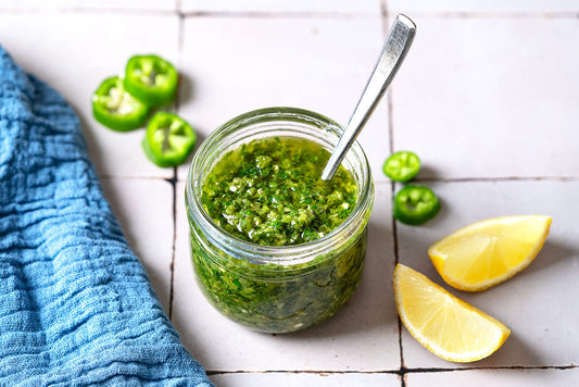 A spoonful of zhoug being held over a jar of the zhoug, with a kitchen towel, sliced jalapenos and lemon wedges in the background.