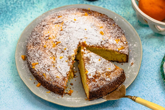 A round orange cardamom olive oil cake with a golden-brown crust, dusted with powdered sugar and sprinkled with fresh orange zest.
