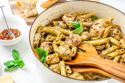 A white bowl filled with pasta in a creamy pesto sauce and chicken with a fresh basil garnish and a bowl of red pepper flakes on the side.
