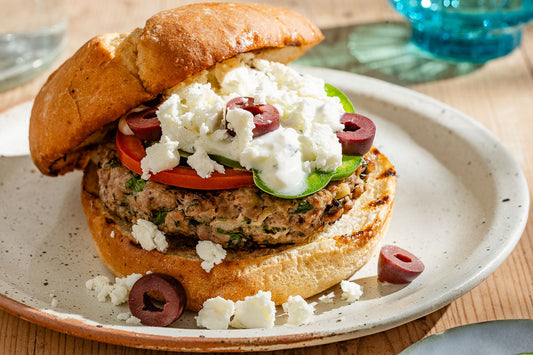 A lamb burger on a plate with a toasted bun topped with feta, vegetables, and olives.