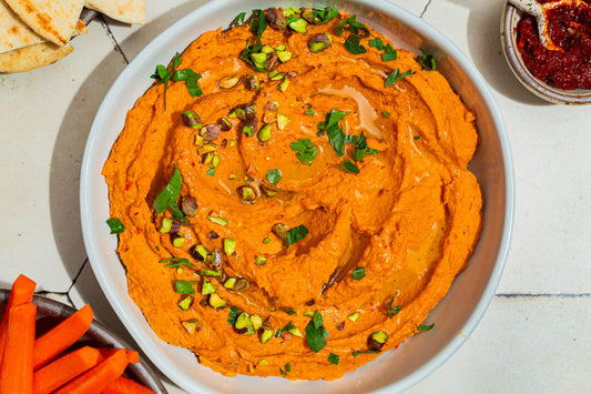 An overhead photo of a large bowl filled with orange hummus made with carrots and topped with pistachios and fresh herbs.