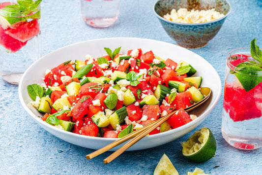 A bowl of chopped watermelon and cucumber garnished with feta mint with two water glasses beside it.