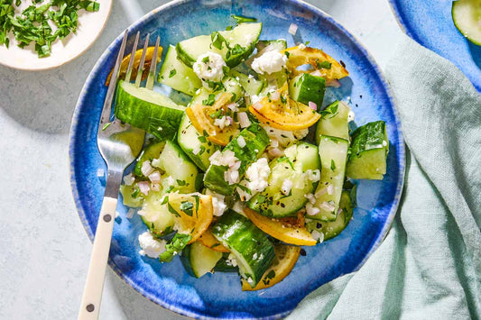 An overhead photo of a blue plate topped with chopped cucumber, lemon, and feta garnished with fresh herbs.