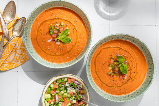 An overhead photo of two bowls filled with watermelon gazpacho soup and a side bowl of fresh chopped vegetables.