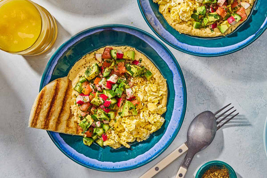 An overhead photo of a blue bowl filled with hummus, chopped vegetables, and pita bread.
