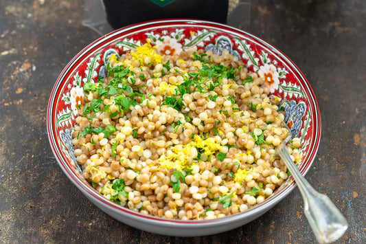 A decorative bowl filled with pearl couscous and topped with fresh lemon zest and herbs.