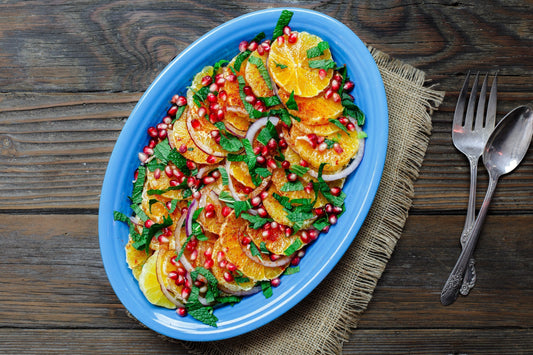 An overhead photo of a blue serving dish topped with sliced oranges, pomegranate seeds, and fresh herbs.