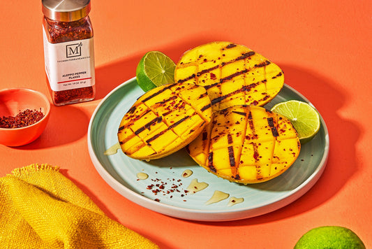 A plate with three halves of grilled mango topped with lime wedges and aleppo pepper seasoning.