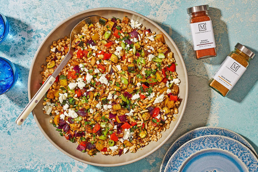 An overhead photo of a bowl with barley, roasted vegetables, and fresh herbs with two bottles of seasoning on the side.