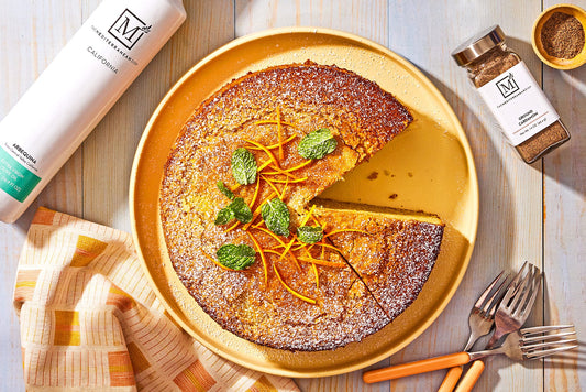 A round orange cardamom olive oil cake with a golden-brown crust, dusted with powdered sugar and sprinkled with fresh orange zest.