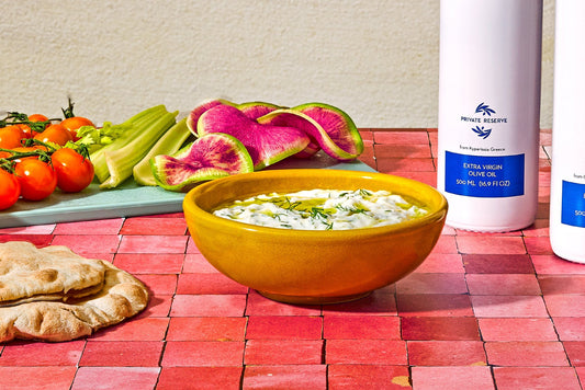 A bowl of tzatziki dip with two olive oil bottles, pita bread, and fresh vegetables in the background.