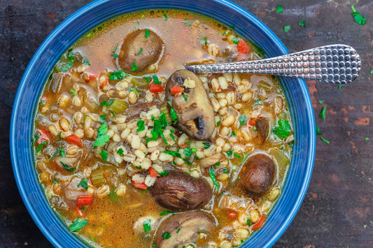 An overhead photo of a bowl with a spoon in it filled with mushroom barley soup and topped with fresh herbs.