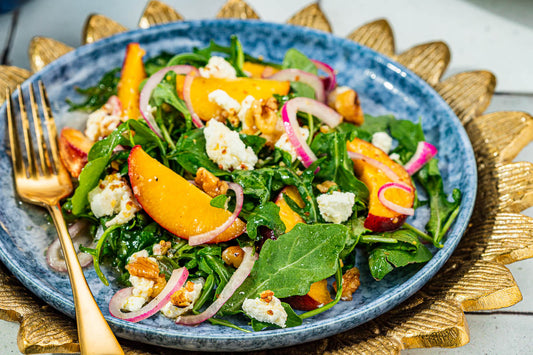 A blue bowl topped with mixed green, sliced peaches, goat cheese, and a gold fork.