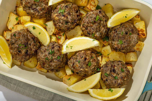 An overhead photo of a casserole dish filled with chopped potatoes, beef meatballs, and lemon wedges.