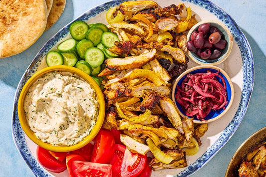 An overhead photo of a large serving plate with chicken shawarma, sliced cucumbers, olives, tomatoes, and a bowl of tzatziki dip.