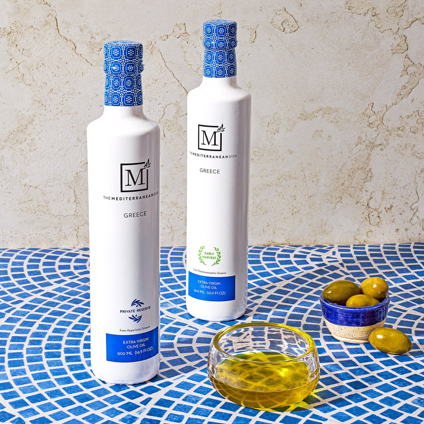 Two bottles of olive oil with decorative blue caps on a textured surface with olives.