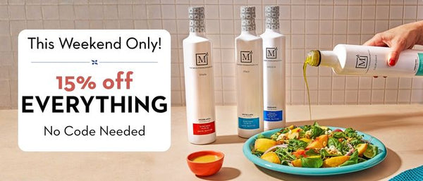 Three white bottles of The Mediterranean Dish olive oils with silver caps and a logo starting with 'M' are displayed on a beige countertop. A hand is pouring olive oil from one of the bottles onto a salad on a blue plate.