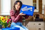 Suzy Karadsheh sitting on a kitchen counter top holding a blue gift box labeled Mediterranean Mystery Box.