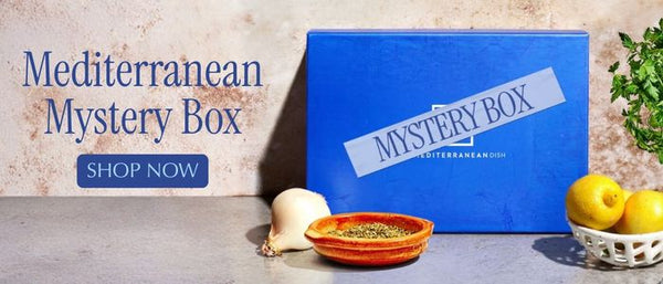 A blue box labeled 'Mediterranean Mystery Box' with a 'SHOP NOW' callout on the left, shown alongside garlic, a small bowl of herbs, lemons, and greenery on a stone surface.