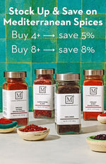 Four glass spice jars from The Mediterranean Dish with white labels and silver lids, arranged on a light surface against a teal tiled background. White promotional text at the top reads, 'Stock Up & Save on Mediterranean Spices' with buy thresholds and savings.