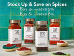 Four glass spice jars from The Mediterranean Dish with white labels and silver lids, arranged on a light surface against a teal tiled background. White promotional text at the top reads, 'Stock Up & Save on Mediterranean Spices' with buy thresholds and savings.