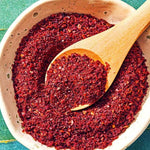 A close-up overhead shot of a light beige speckled bowl filled with coarse ground sumac spice, with a wooden spoon holding a scoop of the powder on a green surface.