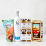 A curated collection of gourmet food products from The Mediterranean Dish arranged on a white surface against a light textured wall: a bag of Food to Live lentils, a tall white bottle of Nocellara olive oil, four glass spice jars with silver lids, and a large bag of Duru extra fine bulgur wheat.