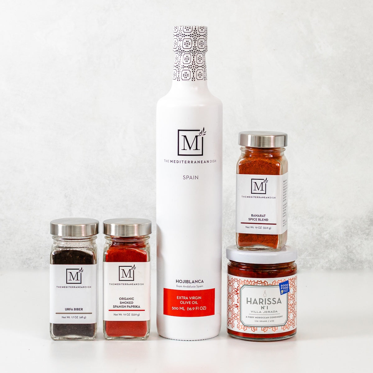 A collection of pantry items from The Mediterranean Dish featuring a white bottle of olive oil, 3 jars of spices, and a jar of Harissa by Villa Jerada.