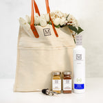A beige canvas tote bag with brown leather handles, filled with white tulips, styled with a white bottle of olive oil, two spice jars, and a metal oil pourer on a light surface against a white background. The bag and bottle feature an 'M' logo from The Mediterranean Dish.