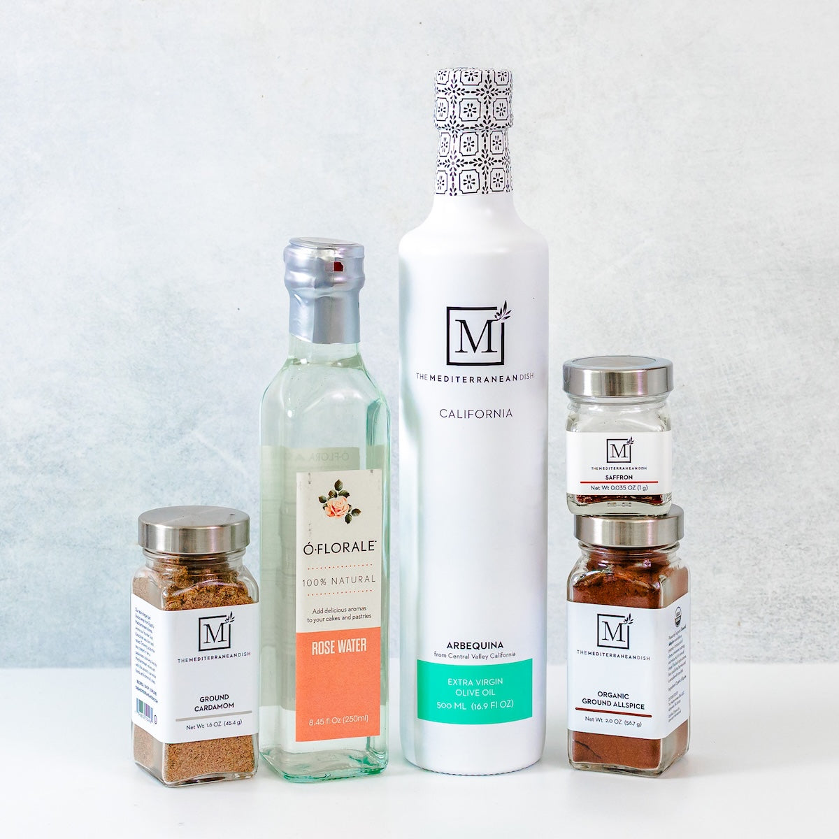 A collection of gourmet food products from The Mediterranean Dish, including a large white bottle of Arbequina olive oil, a clear bottle of Rose Water, and three spice jars, arranged on a white surface against a light gray textured background.