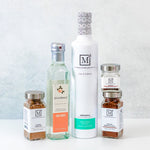 A collection of gourmet food products from The Mediterranean Dish, including a large white bottle of Arbequina olive oil, a clear bottle of Rose Water, and three spice jars, arranged on a white surface against a light gray textured background.