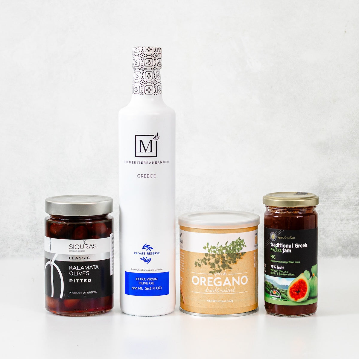 Four gourmet food items arranged on a white surface against a light gray textured background: a tall white bottle of Koroneiki Extra Virgin Olive Oil, a jar of Siouras Classic Kalamata Olives, a can of Oregano, and a jar of Traditional Greek Jam.