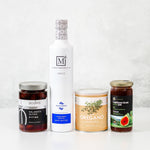 Four gourmet food items arranged on a white surface against a light gray textured background: a tall white bottle of Koroneiki Extra Virgin Olive Oil, a jar of Siouras Classic Kalamata Olives, a can of Oregano, and a jar of Traditional Greek Jam.