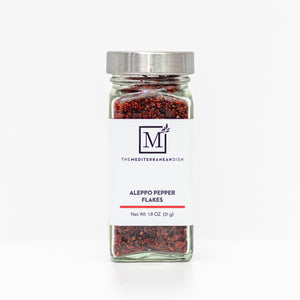 A glass spice jar filled with Aleppo pepper flakes, featuring a white label with the brand logo 'The Mediterranean Dish' and a silver metal lid, set against a clean white background.