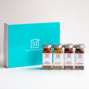 A gift set of four glass spice jars with silver lids lined up in front of a bright turquoise box. The jars have white labels with a black 'M' logo and text including 'MEDITERRANEAN SPICE BLEND', 'RED ORANGE SEEDS', 'KUMIN SEEDS', and 'SAGE'.