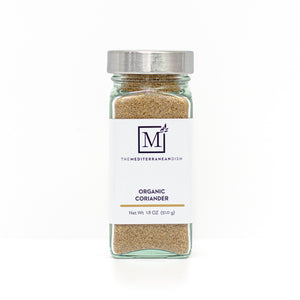 A glass spice jar filled with coriander, featuring a white label with the brand logo 'The Mediterranean Dish' and a silver metal lid, set against a clean white background.