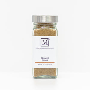 A glass spice jar filled with organic cumin seasoning, featuring a white label with the brand logo 'The Mediterranean Dish' and a silver metal lid, set against a clean white background.